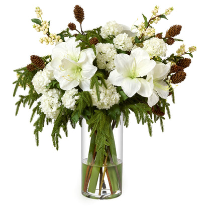 A faux floral arrangement by Diane James Home  featuring large white amaryllis, white snowballs, green pine, brown pinecones, and white snowberries in a clear cylindrical glass vase on a white background.