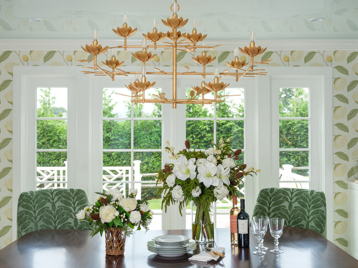 A large multi-light chandelier with a warm gold finish hangs over a dining table in a bright room with white-framed windows, green patterned wallpaper, and green upholstered chairs. Faux floral arrangements by Diane James Home and dining table settings are visible in the foreground.