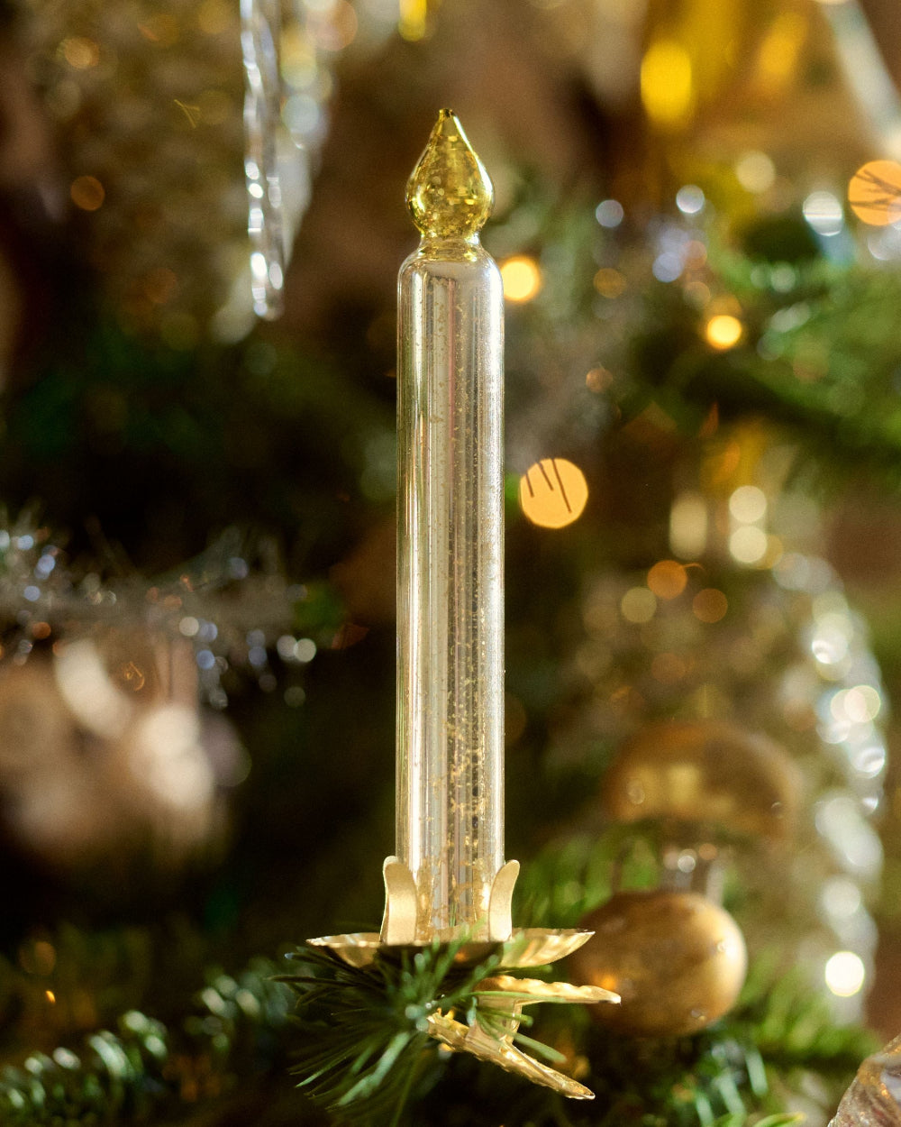 A clear glass taper candle ornament by Eddie Ross with a gold glitter finish, featuring a pointed gold top and a decorative gold base, displayed on a decorated Christmas tree with green branches and warm bokeh lights.