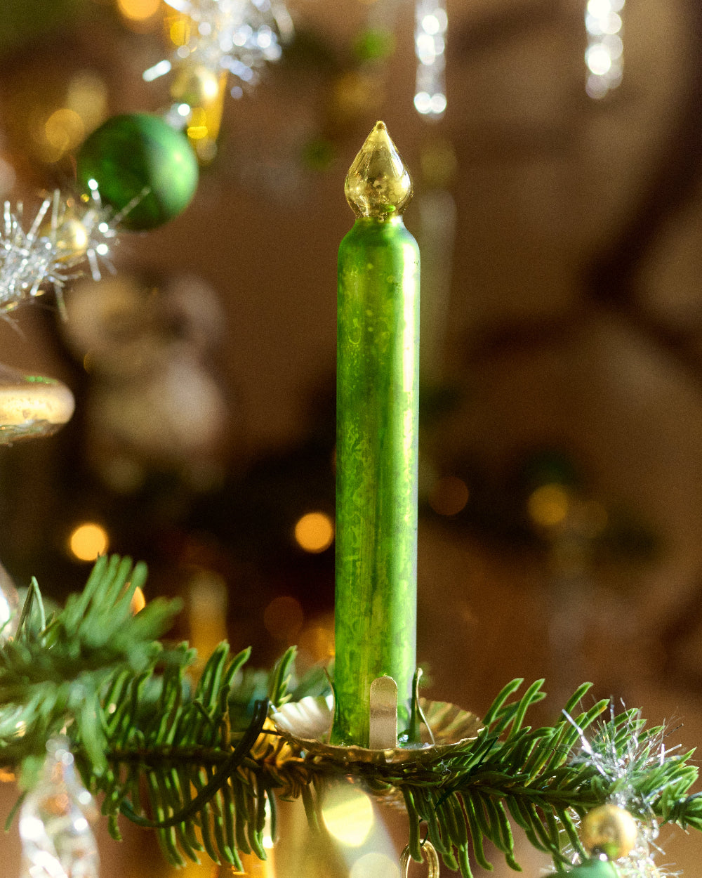 A single green candle-shaped Christmas ornament by Eddie Ross with a metallic gold top, displayed on a green pine branch with holiday decorations in a warm, blurred background.