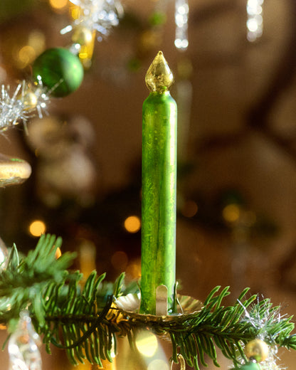 A single green candle-shaped Christmas ornament by Eddie Ross with a metallic gold top, displayed on a green pine branch with holiday decorations in a warm, blurred background.