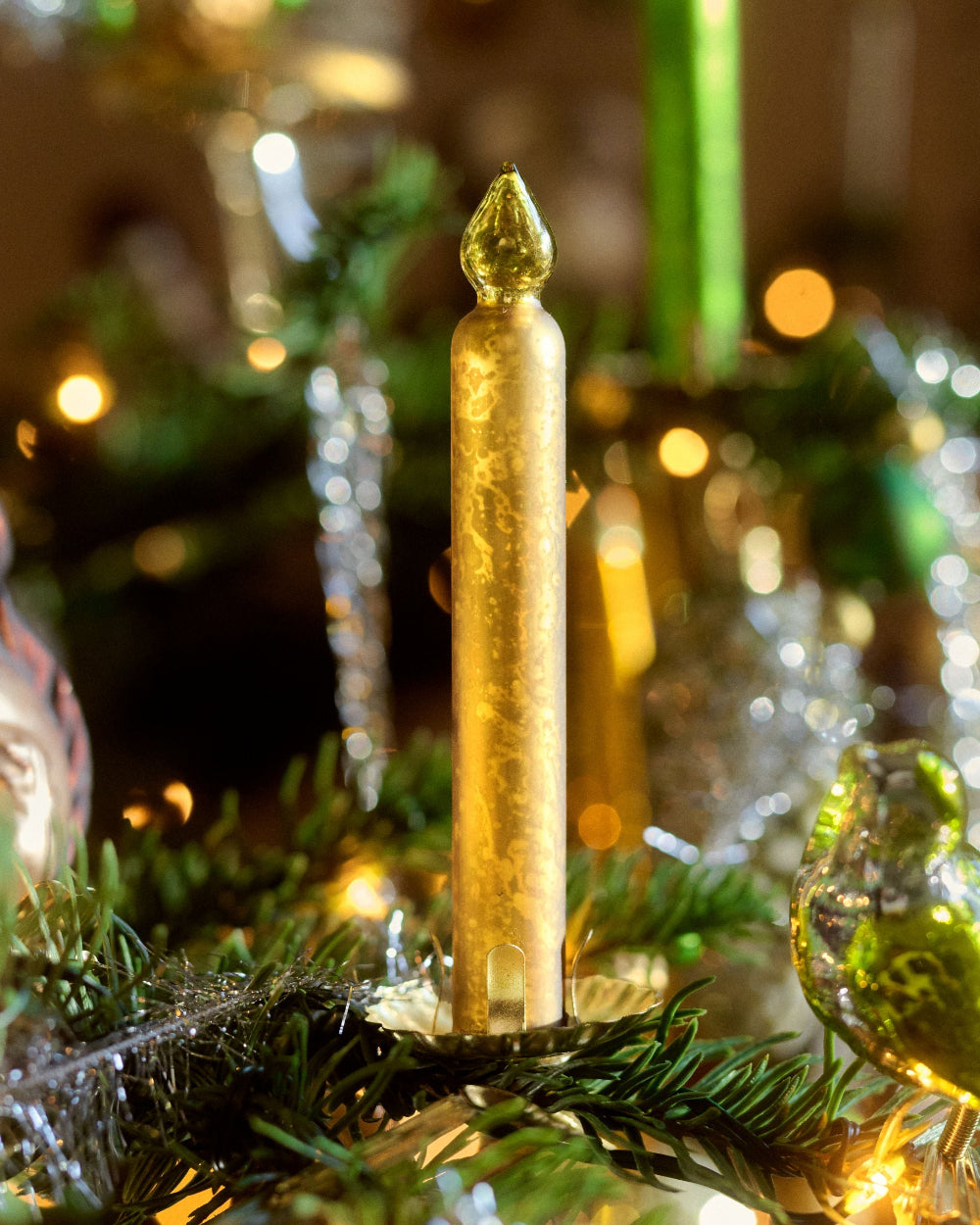A gold Christmas candle ornament by Eddie Ross with a textured, glittery finish standing upright in a pine garland with warm bokeh lights and holiday decorations in the background.