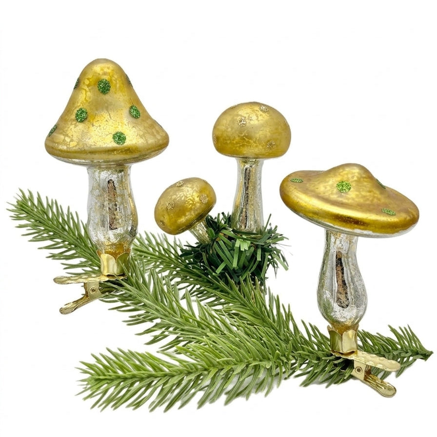 Three gold and silver glass mushrooms with green accents by Eddie Ross, displayed on green pine branches against a white background.