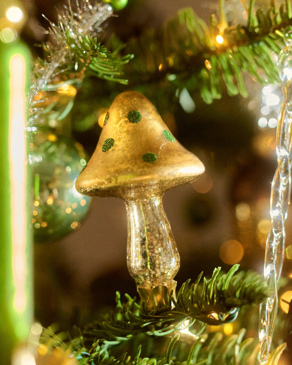 A close-up of a gold mushroom-shaped Christmas ornament with green glittery spots by Eddie Ross, mounted on a green pine tree branch with warm holiday lights in the background.