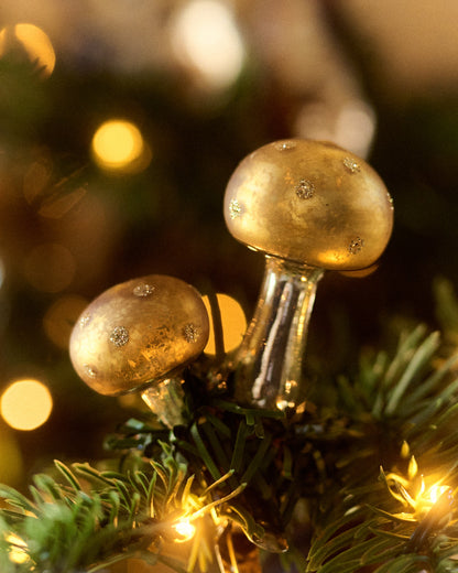 Two decorative gold mushrooms by Eddie Ross attached to green pine branches with warm white string lights and a soft golden bokeh background.