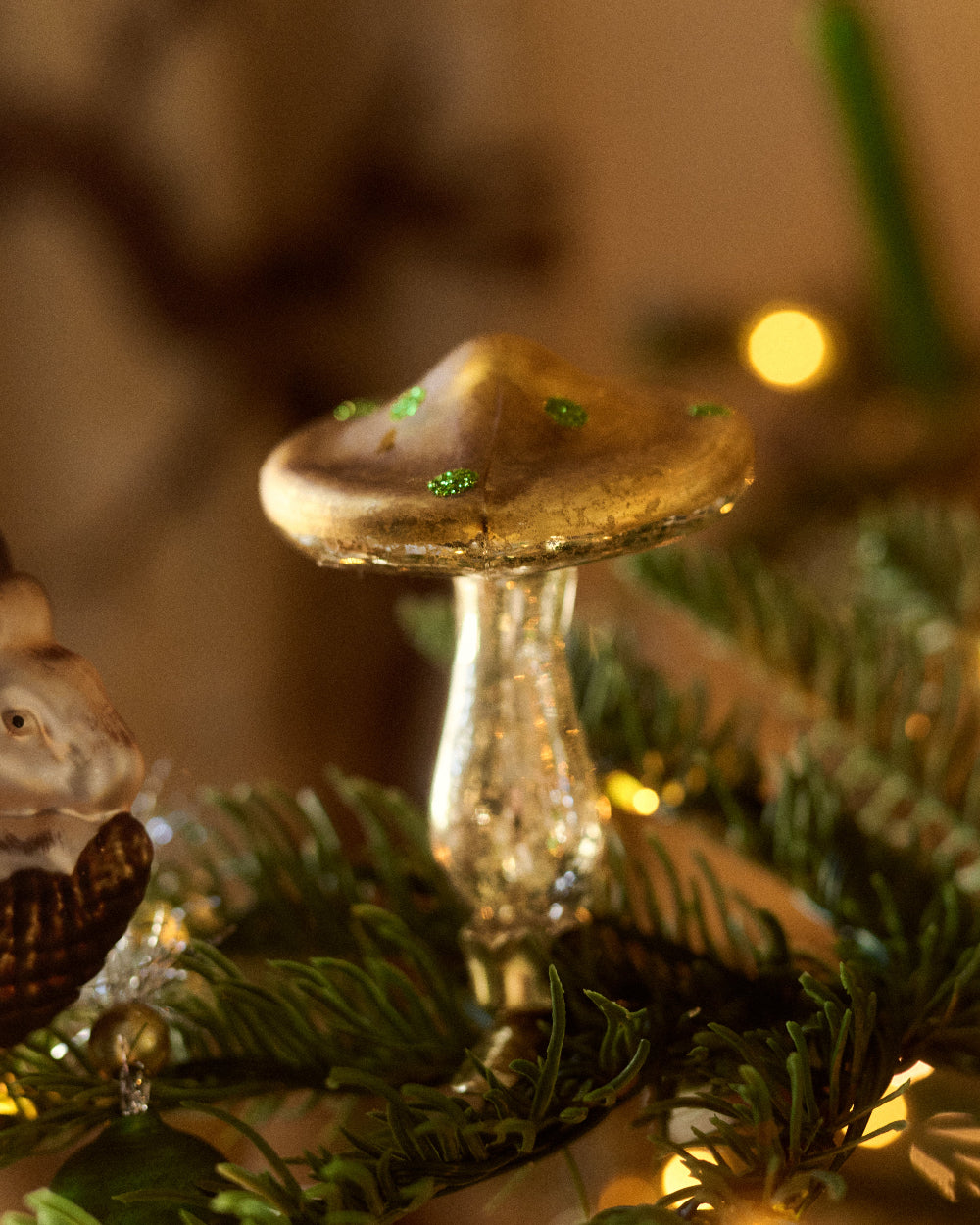 A close-up of a decorative mushroom ornament with a gold cap and glittery stem by Eddie Ross, nestled among green pine branches and warm string lights.